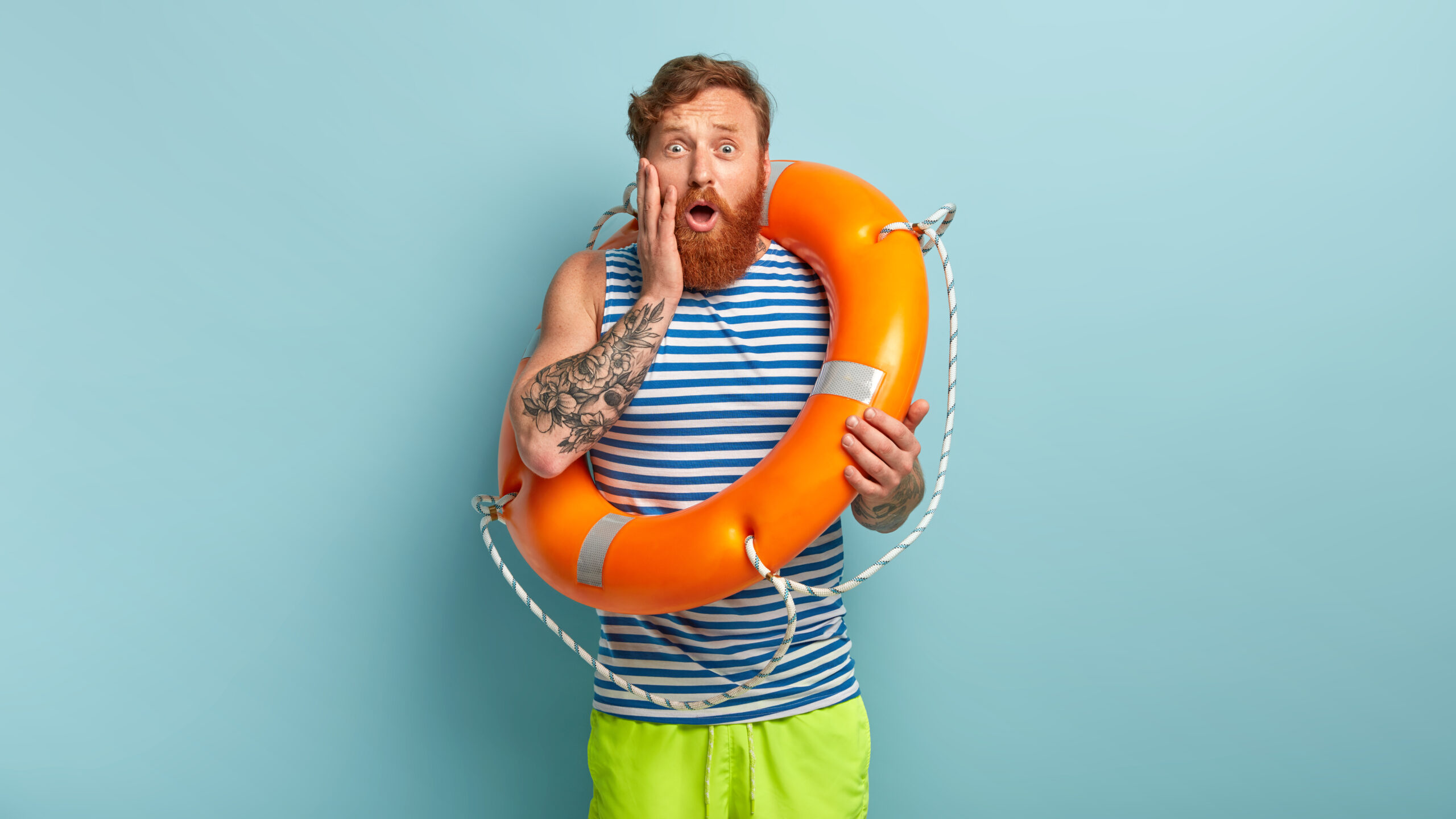 Impressed redhaired man has nervous facial expression, afraids of swimming in ocean for first time, uses ring buoy, wears striped white and blue t shirt, green shorts, cares about safety on water